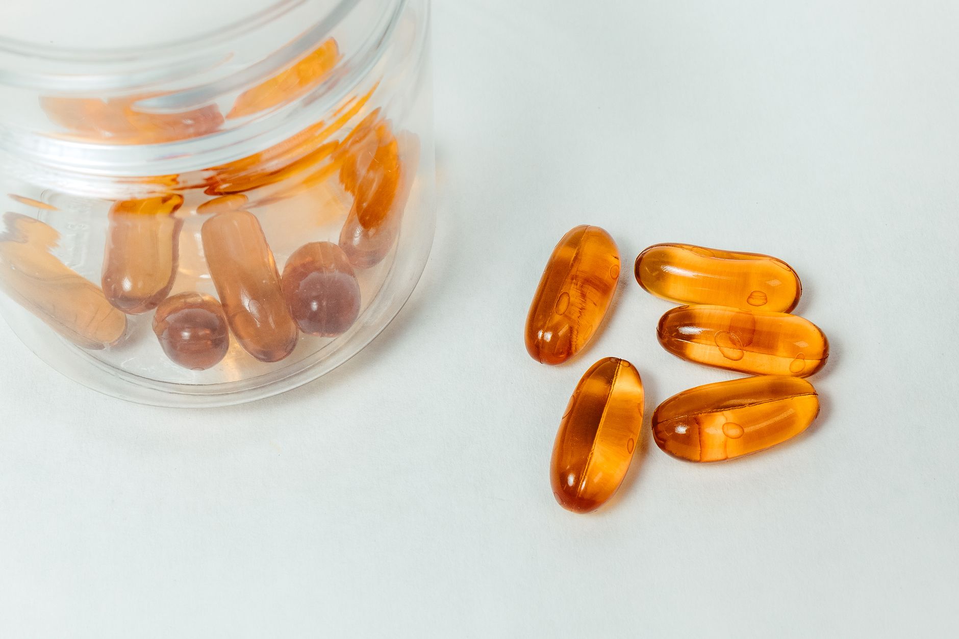 Five amber softgel capsules beside a tipped glass jar of capsules on a white surface